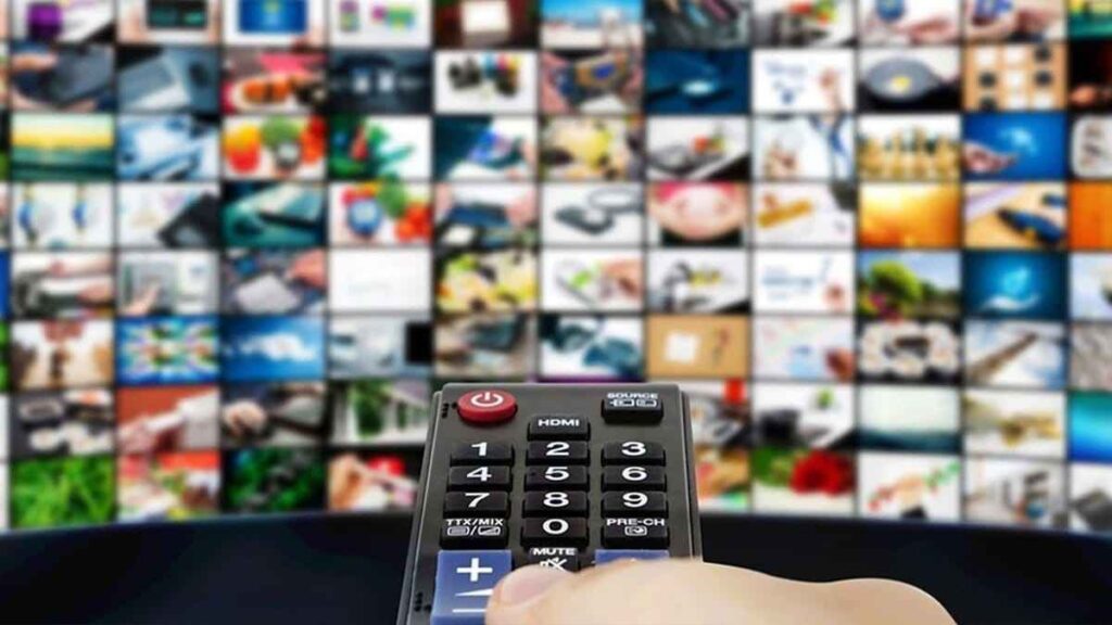 A remote control is held up against a blurred background of a television screen showing a large grid of diverse channels and content thumbnails. [IPTV Not Showing Favorite Channels]